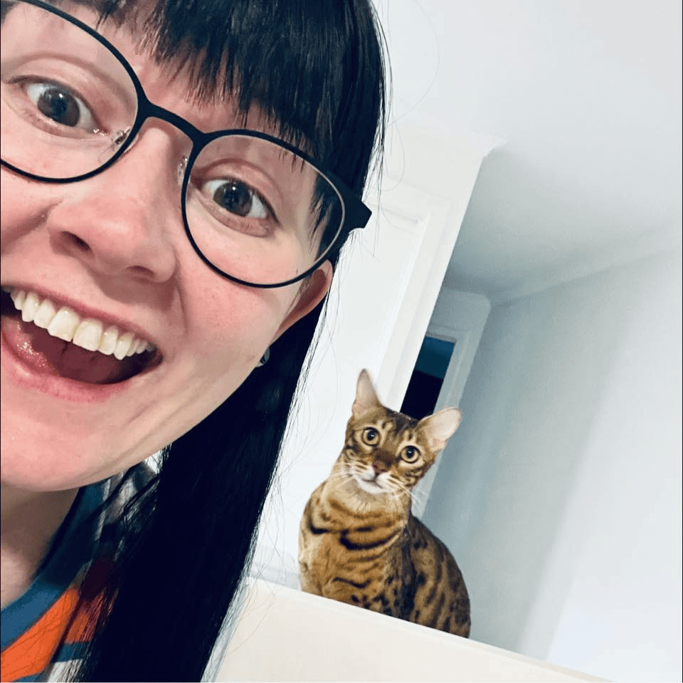 A smiling woman wearing glasses taking a selfie with a Bengal cat sitting behind her on a ledge.