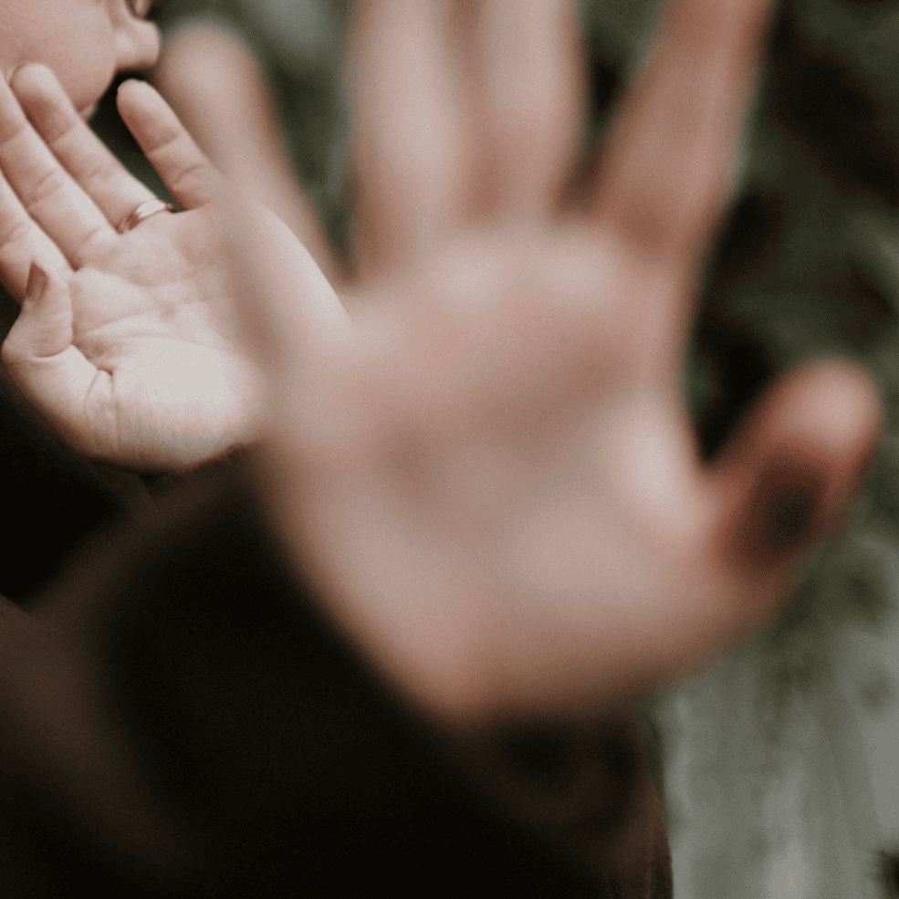 A person holding their hand up toward the camera, partially blocking the lens, with a blurred background.